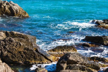 Turkuaz deniz taşı plajı, parlak güneş ışığında dalgalar kırılıyor. Renkli parlak deniz arka planı. Yaz konsepti, tatil, seyahat. Saf temiz deniz suyu, sahilde büyük taşlar.