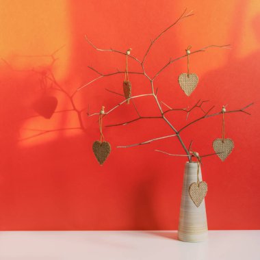 Sevgililer Günü için beyaz ve kırmızı arka planda yaratıcı kompozisyon. Kalpler masanın üzerindeki vazoda bir dalın üzerinde. El yapımı, kaba bir çuval bezi. Sıcak ev atmosferi, romantik tebrik kartı, kare format