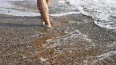 Bir adam deniz kıyısında yürür. Bacaklar yakın. Dalgalar kıyıya vurur ve geçen bir insanı yıkar. Yaz tatili kavramı. Zevk, rahatlama. Kumsalda kum ve küçük çakıl taşları.