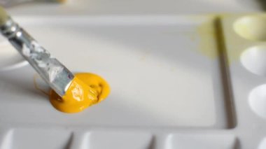 Palette sarı akrilik boya var. Sanatçı elini yavaşça hareket ettirerek boyayı bir fırçayla karıştırır. Yaratıcı süreç, sanat terapisi, ilham, hobi. Ferahlatıcı renkte sıvı akrilik.