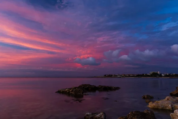 Pink sunset on the sea. Bright juicy landscape with low cumulus clouds on the horizon. Purple, lilac and pink shades of the evening sky. The smooth surface of the sea, the stone shore. The calm idyll