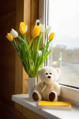 Yellow and white tulips in a white vase on the window with a teddy bear and a book. Vertical composition, greeting card, Mother's Day, International Women's Day, Birthday. Life style. Hello, spring.