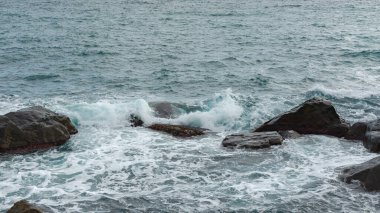 Turkuaz deniz taşı plajı, bulutlu bir bahar gününde dalgalar kırılıyor. Güzel bir deniz arka planı. Yaz konsepti, tatil, seyahat. Saf temiz deniz suyu, sahilde büyük taşlar.