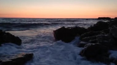 Büyük dalgalar kayaların üzerinde yuvarlanıyor. Okyanus, turuncu günbatımı ve ufkun en alt noktası. Yaz konsepti, sıcak akşam, rahatlama. Alacakaranlıkta güzel sıçramalarla doğal arka plan