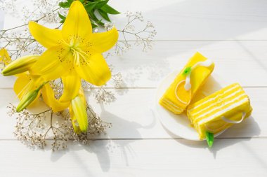 Sarı kek sarı zambaklar beyaz ahşap arka planda parlak güneş ışığında. Tatil için çiçek pastası. Doğum günü konsepti, tebrikler, romantik ruh hali. Aydınlatılmış sarı renk