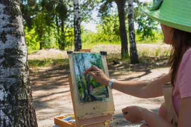 Genç bir kadın plein havası çiziyor. Huş ağacında şapka takan bir sanatçı doğadan bir eskiz yazar. Yaratıcılık konsepti, favori hobi, hobi. Bir resmi akrilik boyalarla boyama süreci