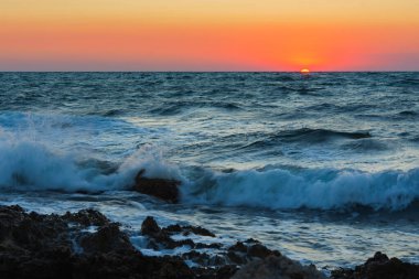 Denizde gün batımı ve fırtına. Batan güneşe karşı büyük dalgalar. Okyanusta bir yaz fırtınası. Kayalık sahilde köpük kırıkları olan güzel bir deniz spreyi. Güzel doğal arka plan. Turuncu batan güneş.