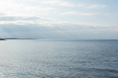 Atmosferik deniz manzarası. Mavi bulutlar parlak güneş ışınları okyanusun pürüzsüz yüzeyini aydınlatır. Ufukta deniz feneri olan panoramik manzara. Barış, sükunet, rahatlama kavramı.