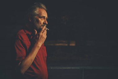 Kırmızı tişörtlü yaşlı adam bir şey bekliyor ve şehir merkezinde sigara içiyor.