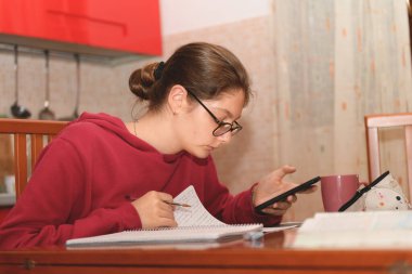 Genç öğrenci mutfağında masasında oturuyor ve ev ödevini yapıyor, o bir cep telefonuyla internete bağlanıyor. Uzaktan öğrenme, evde eğitim ve online eğitim, gerçek hayat anı