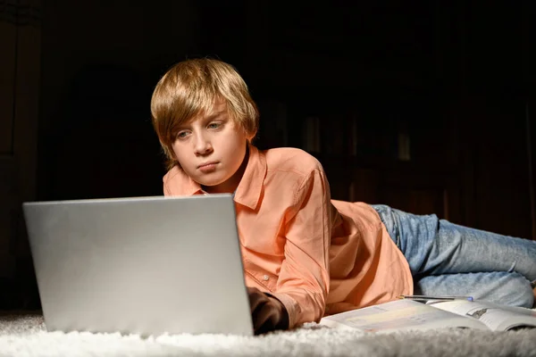 Blond serious boy spending time with notebook, lying on the floor. Homeschooling and education from home online. Computer dependence