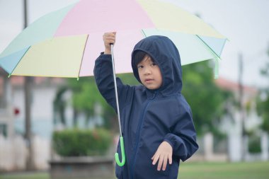Yağmurdan sonra elinde renkli şemsiyeyle parkta oynayan mutlu Asyalı çocuk.