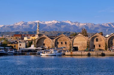 Fishing boats anchored by piers of Old Venetian shipyards or Neoria. Church bell tower and minaret, distant Cretan mountain. Chania, Crete, Greece.