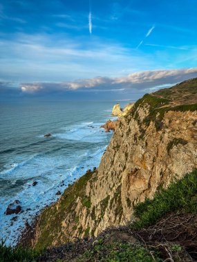 Parlak mavi gökyüzü altında engebeli yüksek uçurumlar ve okyanus dalgalarıyla okyanus manzarası ve güneşli bir günde Cabo da Roca veya Cape Roca, Portekiz, Avrupa 'nın en batı noktası, Ursa sahilinin parlak ışıklı kayaları veya Praia da Ursa. 