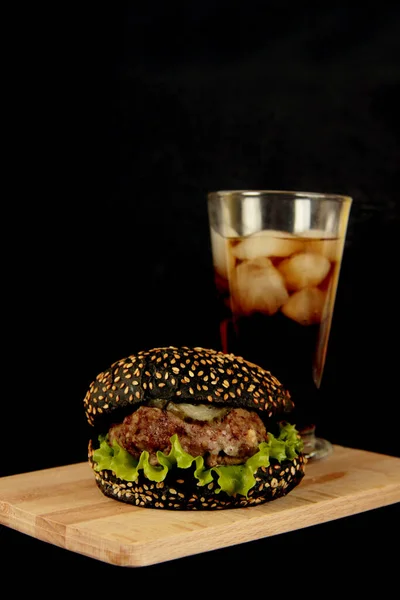 木製のまな板の上に黒いハンバーガーと黒の背景に隔離されたアイスコーラのガラス 画像にはコピースペースがあります — ストック写真