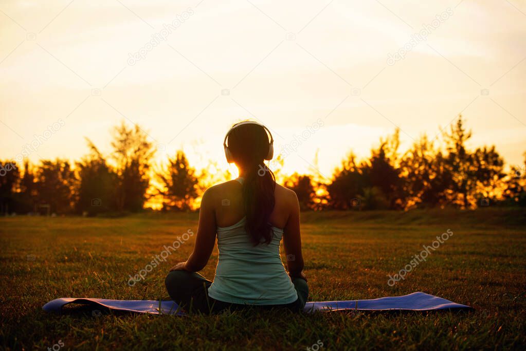 Concept of healthy lifestyle and relaxation-back view of attractive girl in sportswear with headset sitting in lotus position while listening the music outdoor on sunset
