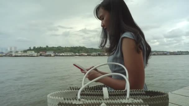 Preciosa Chica Asiática Sentada Orilla Del Río Con Bolsa Junto — Vídeo de stock