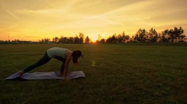 Genç bir kadın gün batımında yeşil parkta yoga egzersizi yapıyor.