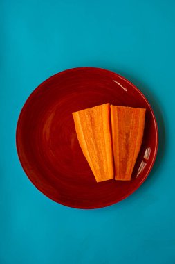 Masanın mavi yüzeyinde kırmızı bir tabakta iki dilim kesilmiş havuç ve bahçe toprağının ışık izleri var. Yaz sezonu. Kapak.