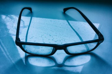 İçerideki kirli mavi bir masanın üstünde siyah çerçeveli gözlük var. Kapak. Web afişi.