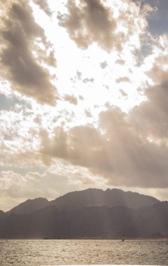 Dağlı manzaralı deniz manzarası ve bulutların arasında parlayan parlak güneş ışınları.