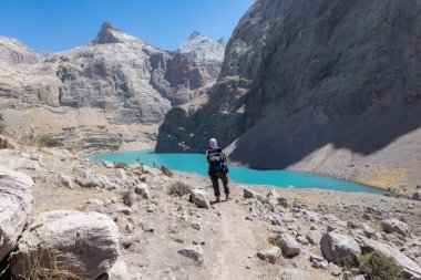 TAJIKISTAN - 20 Ağustos 2025: Dağ sıraları ve turkuaz suyla büyük Allo gölü manzarası ve Fann dağlarında küçük bir yürüyüşçü silueti Pamir Alay Tacikistan