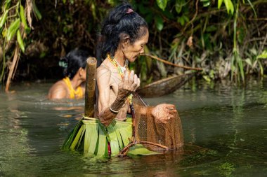 SIBERUT, INDONESIA - 5 Kasım 2025: Mentawai kabilesindeki geleneksel kıyafet ve vücut süslemeleri giyen kadın, Siberut Adası 'ndaki köyünde nehirde balık tutarken fotoğraflandı, Sumatra, Endonezya