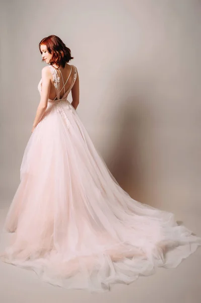 Moda bir gelinlik. Pembe tüllü gelinlikli kızıl gelin ve stüdyosunda uzun bir tren bekliyor. Gri arka planda sırt ayrıntıları olan mor gelinlikli güzel bir gelin. Düğün modası. Arka manzara.