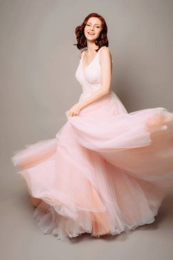 Mutlu gülüşü ve pembe uzun gelinliğiyle stüdyoda dans eden güzel bir gelin. Gelinlik modası. Gri arkaplan ile tam boy portre. 