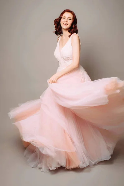 Mutlu gülüşü ve pembe uzun gelinliğiyle stüdyoda dans eden güzel bir gelin. Gelinlik modası. Gri arkaplan ile tam boy portre. 