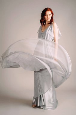 Gri tozlu gece elbisesi giymiş güzel bir kadın. Gri arka planda, yüksek topuklu ayakkabılarla dans eden, gecelikli, kızıl bir kızın stüdyo portresi..