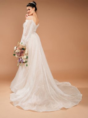 Lüks, ışıltılı, uzun trenli gelinlik. Stüdyoda poz veren genç mutlu gelin. Evlilik kavramı. Pudra renginde moda gelin buketi. Esmer, gülümseyen model.