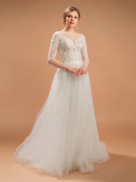 Luxury wedding dress. Fashionable bridal gown with tender french lace and beads, long sleeves, lush tulle skirt. Contemporary design. Beautiful young blonde bride. Studio shot on powder background.