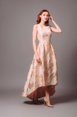 Çiçek desenli, altın şeftali rengi elbiseli kızıl bir kadın. Gri arka planda uzun kolsuz gece elbisesi giyen genç bayanın gizli stüdyo resmi..