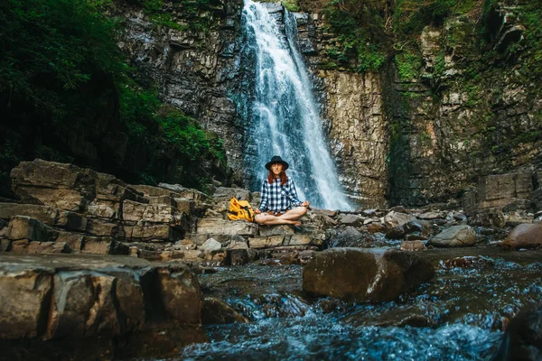 Beautiful girl with red hair in a hat and shirt meditating on rocks in a lotus position against a waterfall. Space for your text message or promotional content — Stock Photo, Image