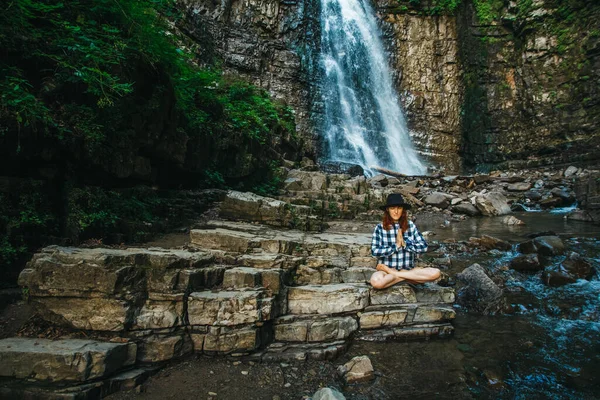 Beautiful girl with red hair in a hat and shirt meditating on rocks in a lotus position against a waterfall. Space for your text message or promotional content — Stock Photo, Image