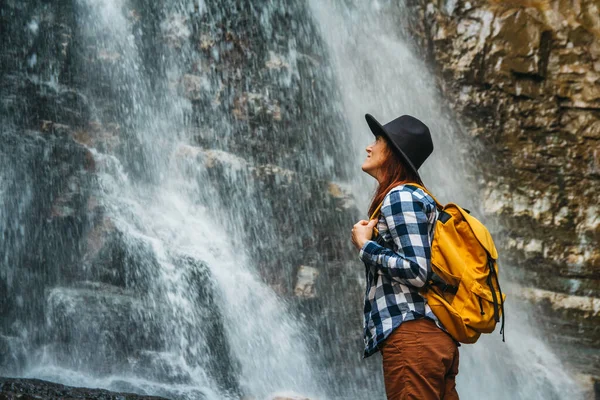 Girl traveler wearing a hat and a yellow backpack while standing against a waterfall. Lifestyle Travel Concept. Space for your text message or promotional content — Stock Photo, Image