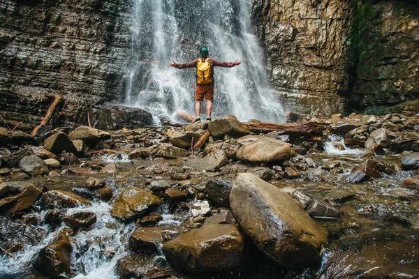 Traveler man with a yellow backpack standing on the background of a waterfall. Hiker couple exploring nature. — Stock Photo, Image