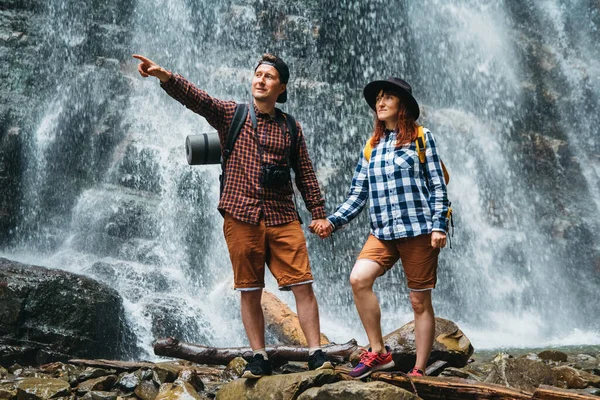 Man and woman hikers trekking a rocky path against the background of a waterfall and rocks. Hiker couple exploring nature. — Stock Photo, Image