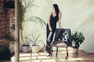 Siyah elbiseli güzel bir manken. Dairenin içinde, koltuğun yanında duruyor..