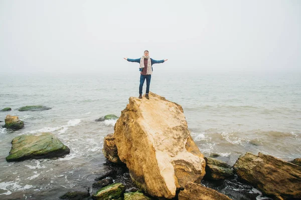 Traveler man stands on a rock against a beautiful sea with waves his hands to the side. Stylish hipster boy posing. Place for text or advertising.