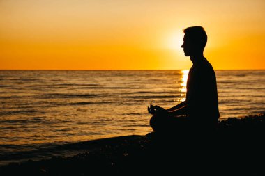 Meditasyon yapan bir adamın silueti sahilde, deniz arka planında ve günbatımında poz verir. Özgürlük rahatlama kavramı. Metin veya reklam için yer.