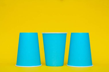 Mavi kağıt bardaklar sarı arka planda sıralanmış. Kopyala, metin için boşluk.