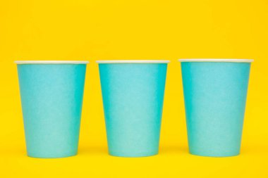 Mavi kağıt bardaklar sarı arka planda sıralanmış. Kopyala, metin için boşluk.