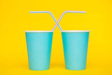 Sarı arka planda renkli plastik pipetler içen iki mavi kağıt bardak. Partiye hazırız. Minimalist stil. Kopyala, metin için boşluk.