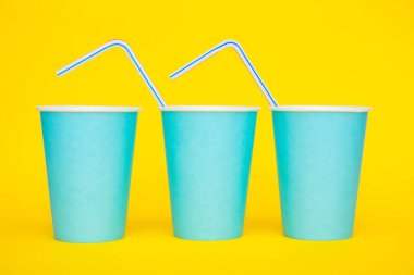 Sarı arka planda renkli plastik pipetler içen mavi kağıt bardaklar. Partiye hazırız. Minimalist stil. Kopyala, metin için boşluk.