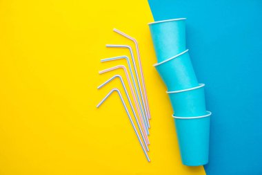 Mavi kağıt bardaklar, renkli plastik pipetler, sarı ve mavi arka plan. Partiye hazırız. Üst Manzara. Minimalist stil. Kopyala, metin için boşluk.