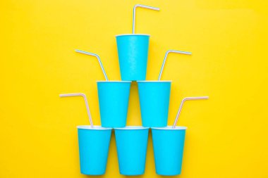 Sarı arka planda renkli plastik pipetler içen mavi kağıt bardaklar piramidi. Partiye hazırız. Üst Manzara. Minimalist stil. Kopyala, metin için boş boşluk.