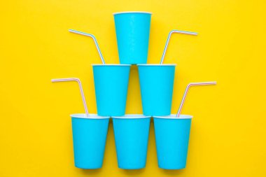 Sarı arka planda renkli plastik pipetler içen mavi kağıt bardaklar piramidi. Partiye hazırız. Üst Manzara. Minimalist stil. Kopyala, metin için boş boşluk.