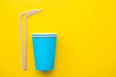 Sarı arka planda renkli plastik pipetler içen mavi kağıt bardaklar. Partiye hazırız. Üst Manzara. Minimalist stil. Kopyala, metin için boşluk.